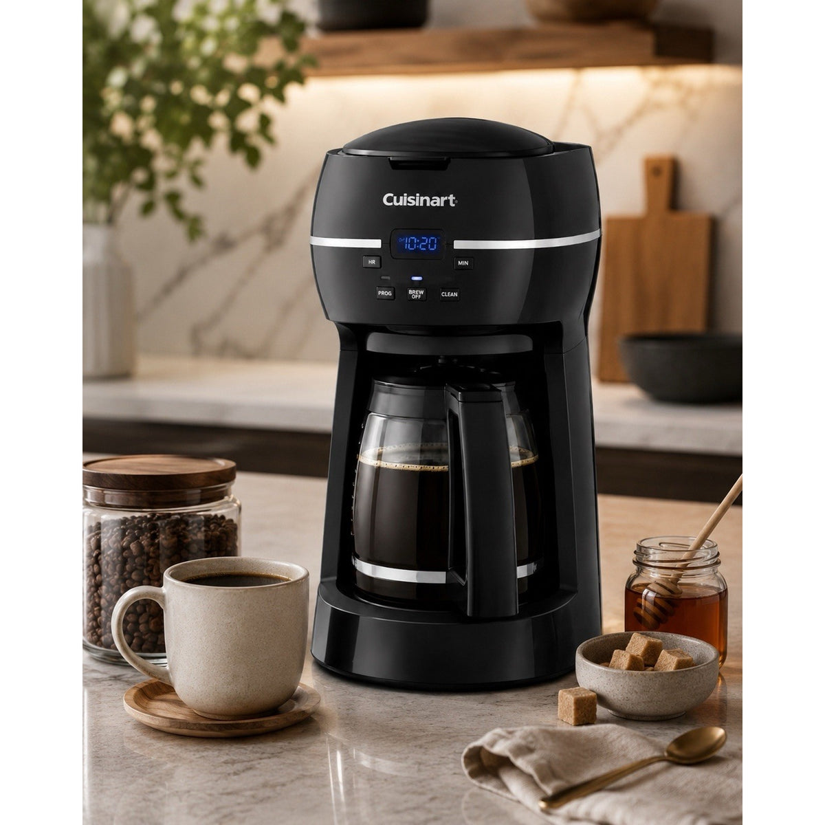 Catalog scene of black Cuisinart DCC-1500FR coffee maker with filled glass carafe, coffee cup, beans, honey, and sugar on a kitchen counter.
